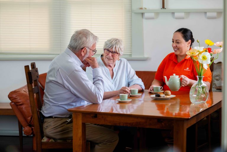 A Helping Hand Care worker in bright orange top sits at a table drinking tea with an older couple, Richard and Jolanta