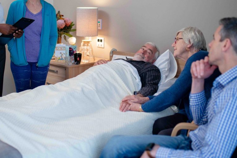 Family gathered round an older man in a bed in an aged care home