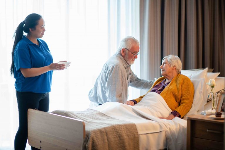 A woman is in a nursing home bed and a man sits at her side. A care worker looks on with a bowl of food for the woman.