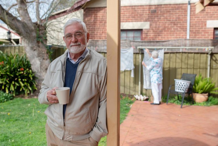 An older man, Richard, looks down the barrel of the camera with his wife struggling to hang out the washing in the background