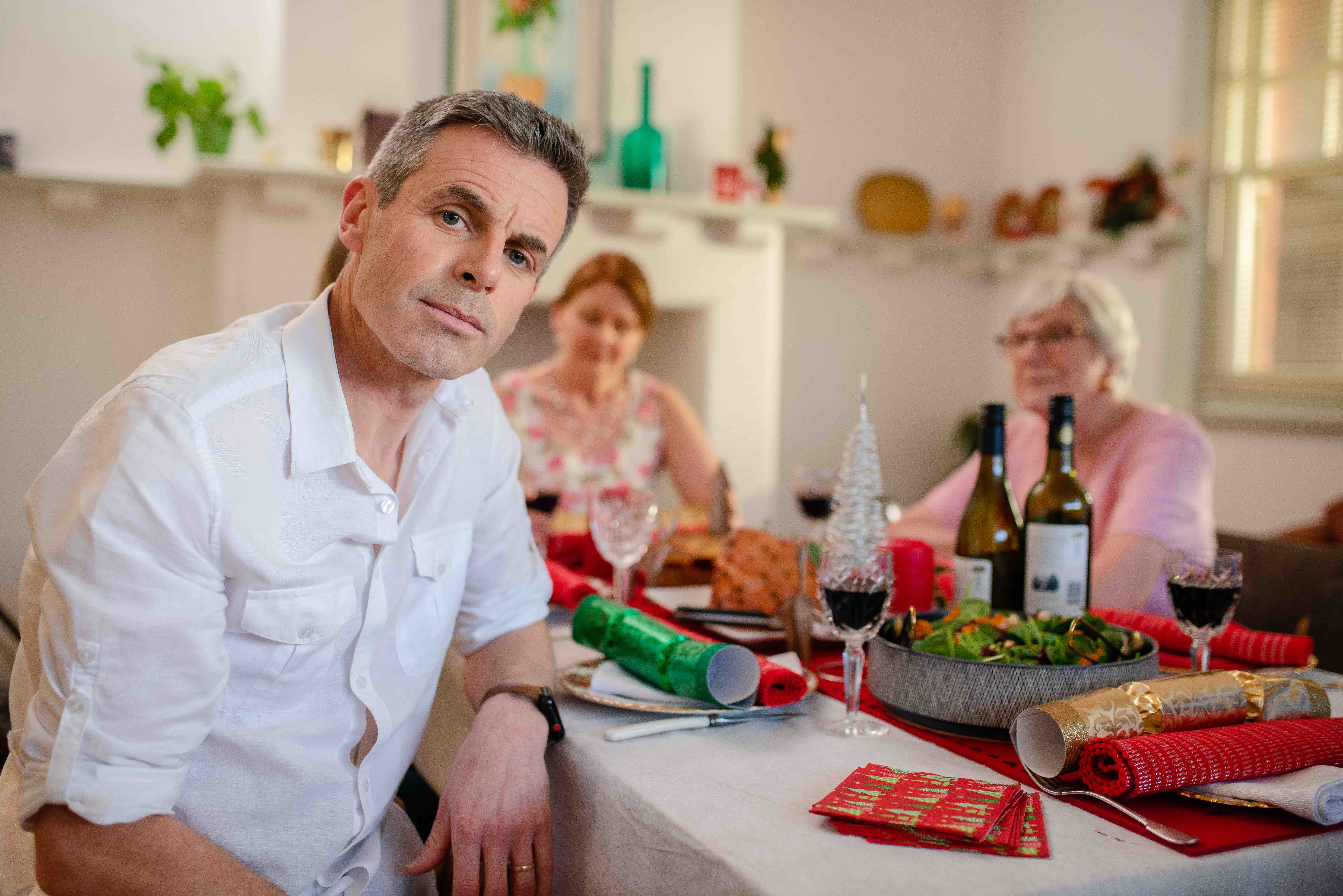 A man looks at the camera, while his sister and parents are in the background