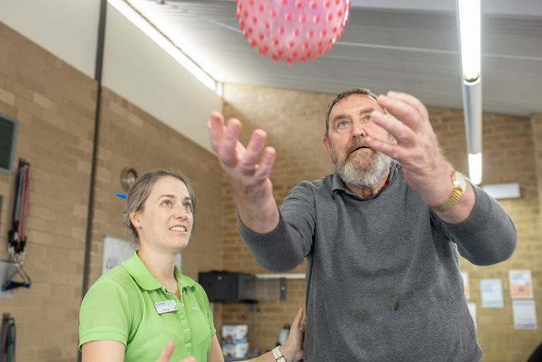 therapy assistant watches on as an older man throws a pink ball